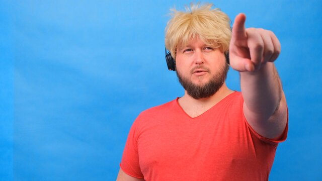freaky fat man in a wig and a pink T-shirt in headphones shows a finger on the camera at you against a blue background.