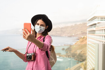Young travel woman taking selfie with mobile smartphone wearing face surgical mask - Travel influencer having fun in vacation during corona virus outbreak - Millennial people and technology concept - obrazy, fototapety, plakaty