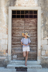 Fototapeta na wymiar Beautiful young female tourist woman standing in front of vinatage wooden door and textured stone wall at old Mediterranean town, smiling, holding, smart phone to network on vacationes.