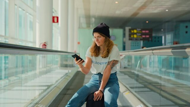 Trendy and fashionable young woman sit on suitcase and pose for selfie photo on social media in big empty airport. Travel blogger or influencer concept. Tshirt design says Cycling in Cyrillic