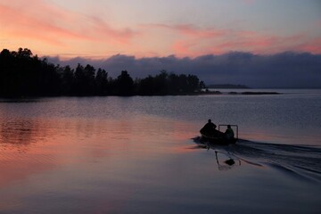 Silhouette of fishermen on the boat in the sunset time. The water is calm but the boat creates small waves. 