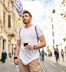 The man walking in the crowded street with his coffee in his hand is wearing a white T-shirt and canvas pants.  man walking with his coffee in his hand on istiklal street