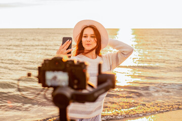 Young woman touching hat and taking selfie in front of camera while standing on beach near waving sea during sunset. Selective focus.