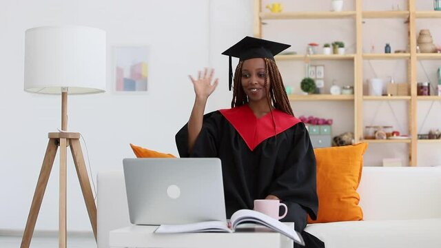 black woman in cap and gown waving, greeting person in video call Spbi. distant graduation from college. technology and academic concept. usa university clothing. use laptop