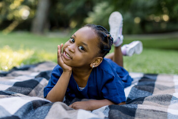 Outdoor portrait of a cute little African girl lying down on the checkered blanket in the park and...