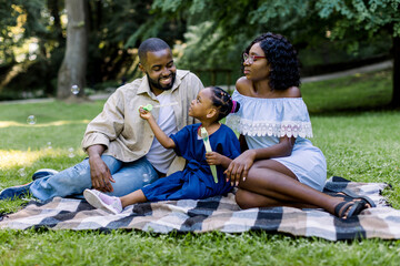Happy young African family playing with soap bubbles with their cute little daughter in park outdoors in summer or spring, sitting on the blanket. Family day concept