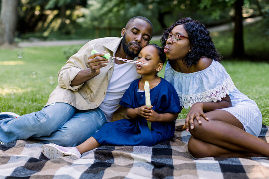Family time, vacations, leisure together. Joyful African American famile with little kid girl blowing bubbles together, having fun on a picnic at city park, sitting on checkered blanket