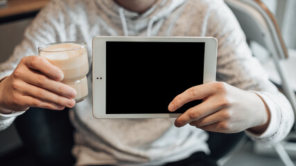 Close up shot of man hold glass cup of coffee and white tablet pc with mock up. The guy is preparing for the remote working day. Copy space. Student sitting in cosy room before distance learning.