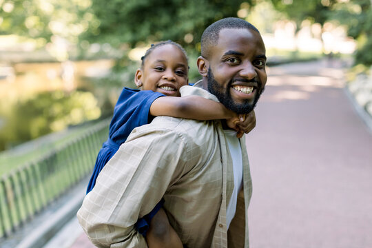 Handsome young dark skinned father and his laughing little daughter, playing in the park. African father piggyback his little daughter outside in nature. Close up portrait