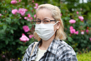 Middle aged mixed race blonde woman with eyeglasses wearing white surgical mask. Protection against coronavirus (COVID-19) and other infectious diseases. Outdoors. Looking at the camera.