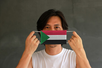 A man with Sudan flag on hygienic mask in his hand and lifted up the front face on dark grey background. Tiny Particle or virus corona or Covid 19 protection.