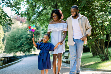 Happy family, joyful time together outdoors. Dark skinned little girl in blue dress, blowing colorful windmill toy, while walking in park alley with her young happy parents on summer day