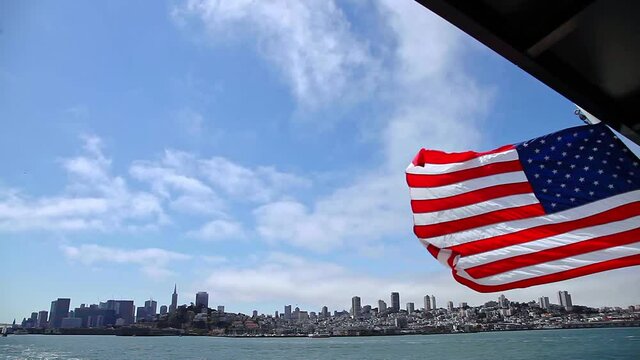 American flag in San Francisco Bay cityscape. Tour to Alcatraz island by ferry boat waving in San Francisco pier with cityscape. Cloudy sky. San Francisco, California, United States