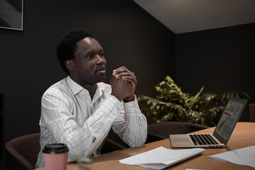 Attractive successful young black businesswoman sitting in his stylish office with open laptop, coffee in disposable cup and papaers on desk, clasping hands, having thoughtful facial expression