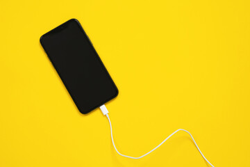 Smartphone is facing up, usb lightning cable or wired headphones are connected to mobile phone connector on yellow background, top view, copy space for advertising text.