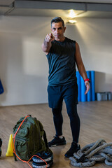 Gym instructor pointing at camera. Motivated boot camp instructor stands with gym equipment in gym...
