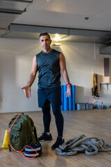 Motivated boot camp instructor stands with gym equipment in gym hall. Dumbbells, rope, sandbag on...