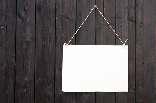 A White Empty Advertising Sign Hangs On A String On A Wall Of Black Boards Close Up