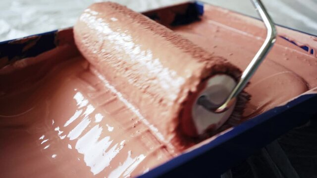 person moves metal roller with brown paint in special blue tray on plastic background before colouring wall in new apartment extreme close view