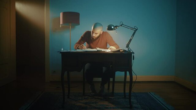 Man writing and searching for notes on a desk with a lamp