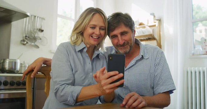 A happy mature couple is making a selfie or video technology call to their relatives with a smart phone in a kitchen at home.
