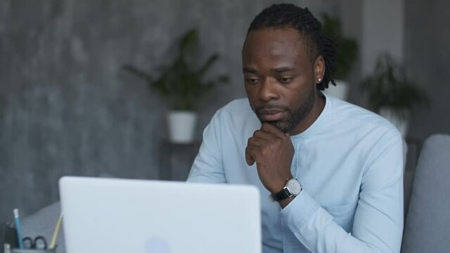 Online education, distance training, business meeting, webinar conference. Portrait of a thinking African American man sitting on a sofa and looking at a laptop