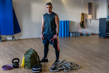 Motivated boot camp instructor stands with gym equipment in gym hall. Dumbbells, rope, sandbag on...