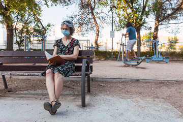 Woman with face mask reading sitting in urban park with a young man near by doing sports, keeping social distance to avoid the spread of the coronavirus in Spain. Selective focus.