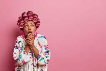 Women and hairstyling concept. Scared housewife looks above with bated breath, applies hair curlers, dressed in casual robe, poses against rosy background with copy space for your advertisement