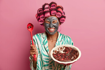 Joyful housewife prepared very tasty chocolate pie, holds big spoon, enjoys eating sweet dessert, smiles broadly, closes eyes, applies beauty mask for reducing wrinkles, wears hair rollers, silk gown