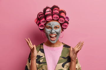 Headshot of angry woman gestures and screams loudly, fed up of beauty procedures, wears hair rollers and cosmetic mask on face, expresses negative emotions. Annoyed houswife shouts at husband