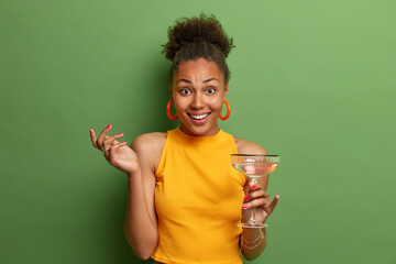 Positive good looking ethnic female poses with glass of summer cocktail, has happy look and nice...
