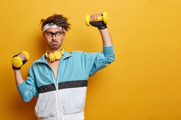 Sporty athletic strong man lifts dumbbells and works hard on training biceps, leads active healthy lifestyle, has regular physical exercises, poses against yellow background, empty space aside