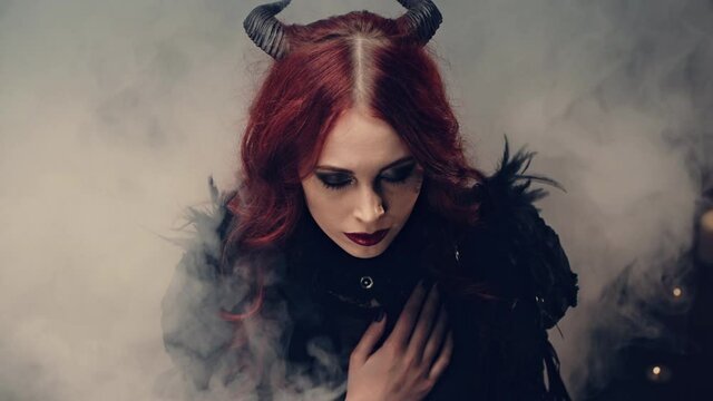 Scary demon doing witchcraft. Red-haired girl witch with black horns sitting on floor in candlelight and praying with folded hands. Halloween mystery rite. Ritual of Occultism or esoterics concept.