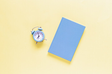 Closed blue notebook and clock on yellow background. Top view, mockup