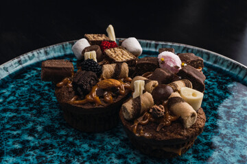 muffins stuffed with delicacy and decorated with chocolates and different sweets on blue plate with black background