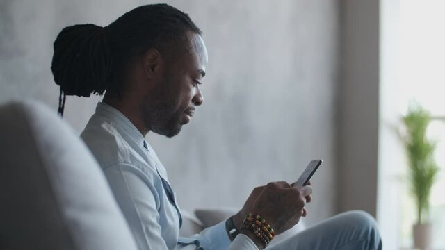 Online education, distance training, business meeting, webinar conference. African American man uses a smartphone while sitting on a sofa near the window