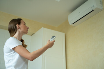 Woman in her modern apartment in living room. She turns on the air conditioner from the remote control. Climate control at home with split system.