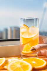hand holds a refreshing drink with ice and slices of orange against the background of the city