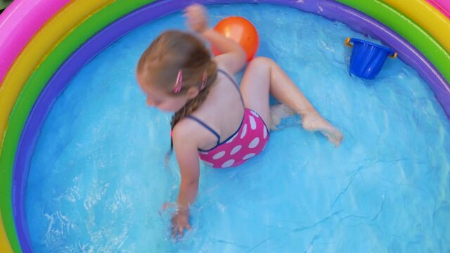 Young girl in pink swimsuit playing in colorful rainbow inflatable swimming pool on a backyard. Summer domestic active lifestyle swim for kids, fun home water leisure for children,hot weather 4K video