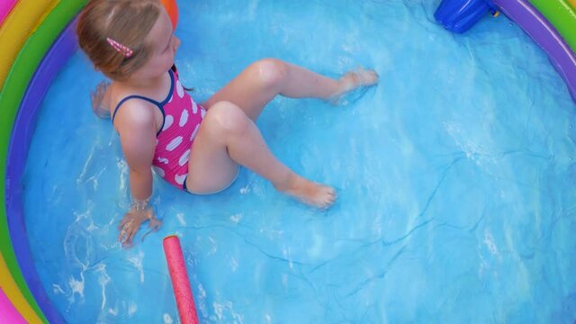 Young girl in pink swimsuit playing in colorful rainbow inflatable swimming pool on a backyard. Summer domestic active lifestyle swim for kids, fun home water leisure for children,hot weather 4K video