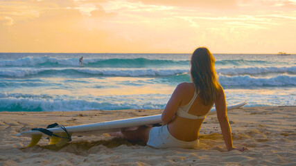 CLOSE UP: Female tourist sits on the sandy beach and watches surfers at sunset