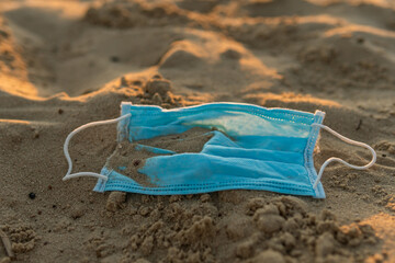 A used disposable mask (protect from COVID-19, Coronavirus) is on a beach, covered with sand, leading to bad consequences like pollution or contamination of the nature and water