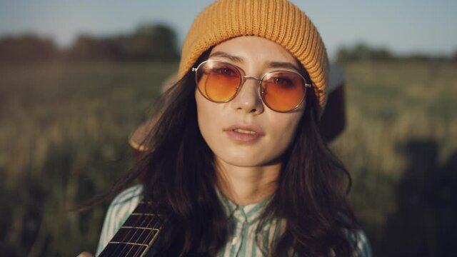 Beautiful Asian hipster woman gutar player walking on a sunny field with a acoustic. The girl in trendy sunglasses enjoy summer sunset and walking forward in countryside. Youth and creativity concept