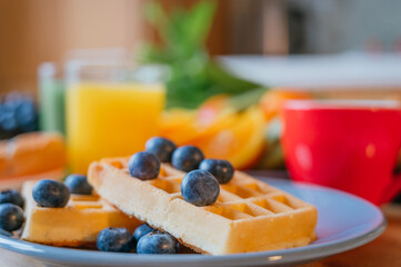 Desayuno saludable de waffles con moras, jugo de naranja, café, croissant, fruta fresaca y jugo verde sobre una mesa de madera