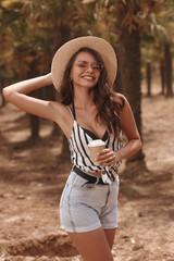 Happy smiling joyous girl. Elegant casual dressed woman with cup of coffee. Stylish female model in shirt, shorts, salty hat and sunglasses standing and posing between palms.