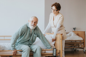Helpful nurse in a beige uniform helps the patient in a blue pajamas getting up from bed