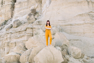 Full length horizontal summer portrait of a young brunette charming woman in fashionable yellow overalls, posing in sand quarry, standing on the stone. Summer fashion