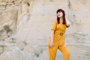 Summer sunny portrait of attractive brunette girl in fashionable yellow clothes overalls posing on the background of desert canyon or sand quarry