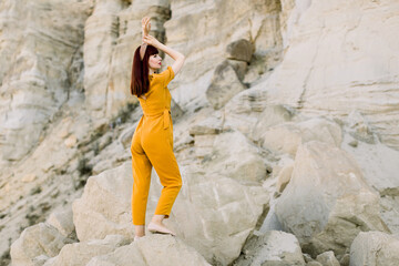 Back angle view of young fashionable lady wearing stylish yellow overalls, with hair hoop band on head, posing in a quarry, in desert or canyon. Fashion lifestyle, modern youth. Copy space